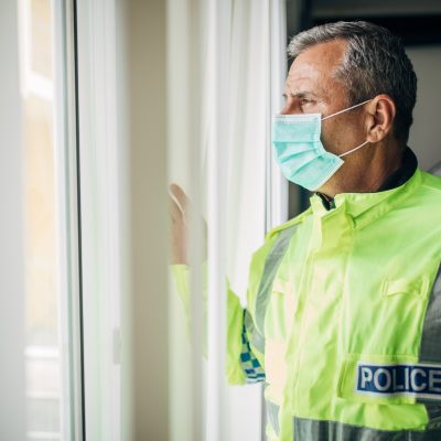 Senior British police officer with protective mask looking through the window from the office. Fight against coronavirus.